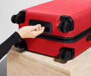 Red suitcase with a hand reaching to open it on a light wooden surface.