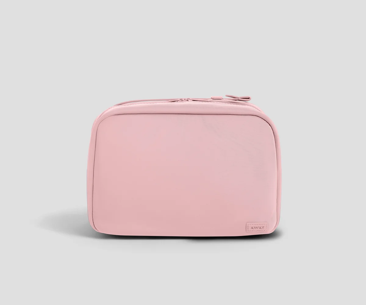 A light pink zippered toiletry bag with rounded corners and a small rectangular "Away" brand tag on the bottom right, shown against a solid light gray background.