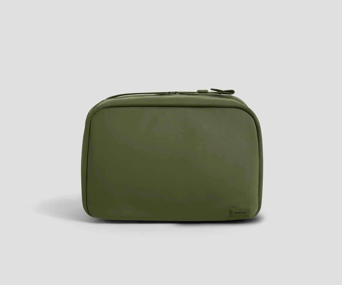 An olive green zippered toiletry bag with a small dark brand tag on the bottom right corner, photographed on a neutral light gray background.