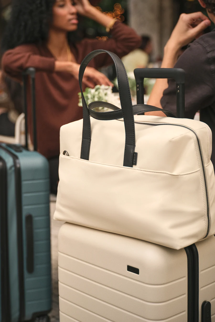 Beige suitcase with black handle and straps, surrounded by people and other suitcases.