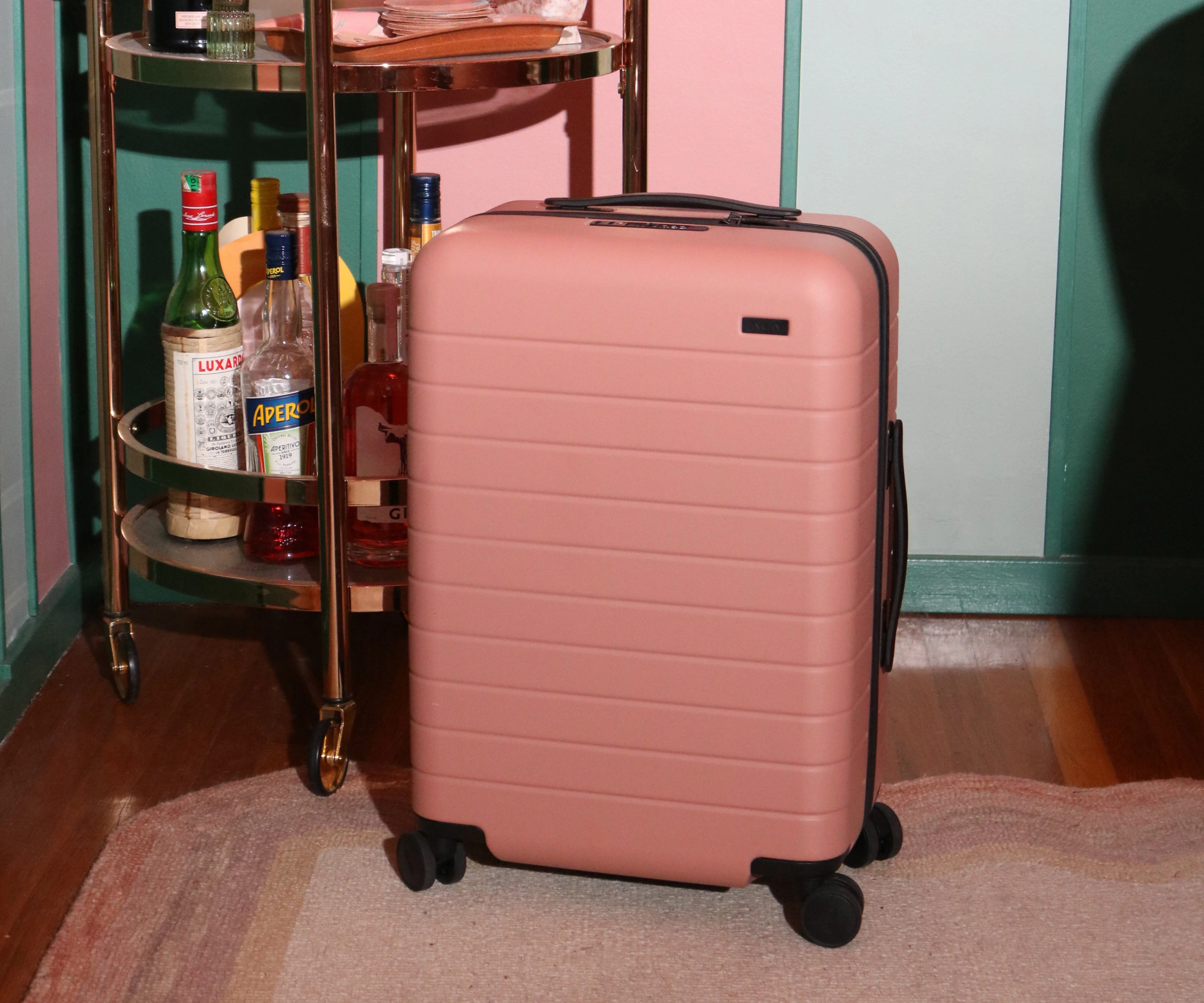 The Carry-On suitcase in Clay Pink shown against a retro room backdrop with a gold bar cart behind it.